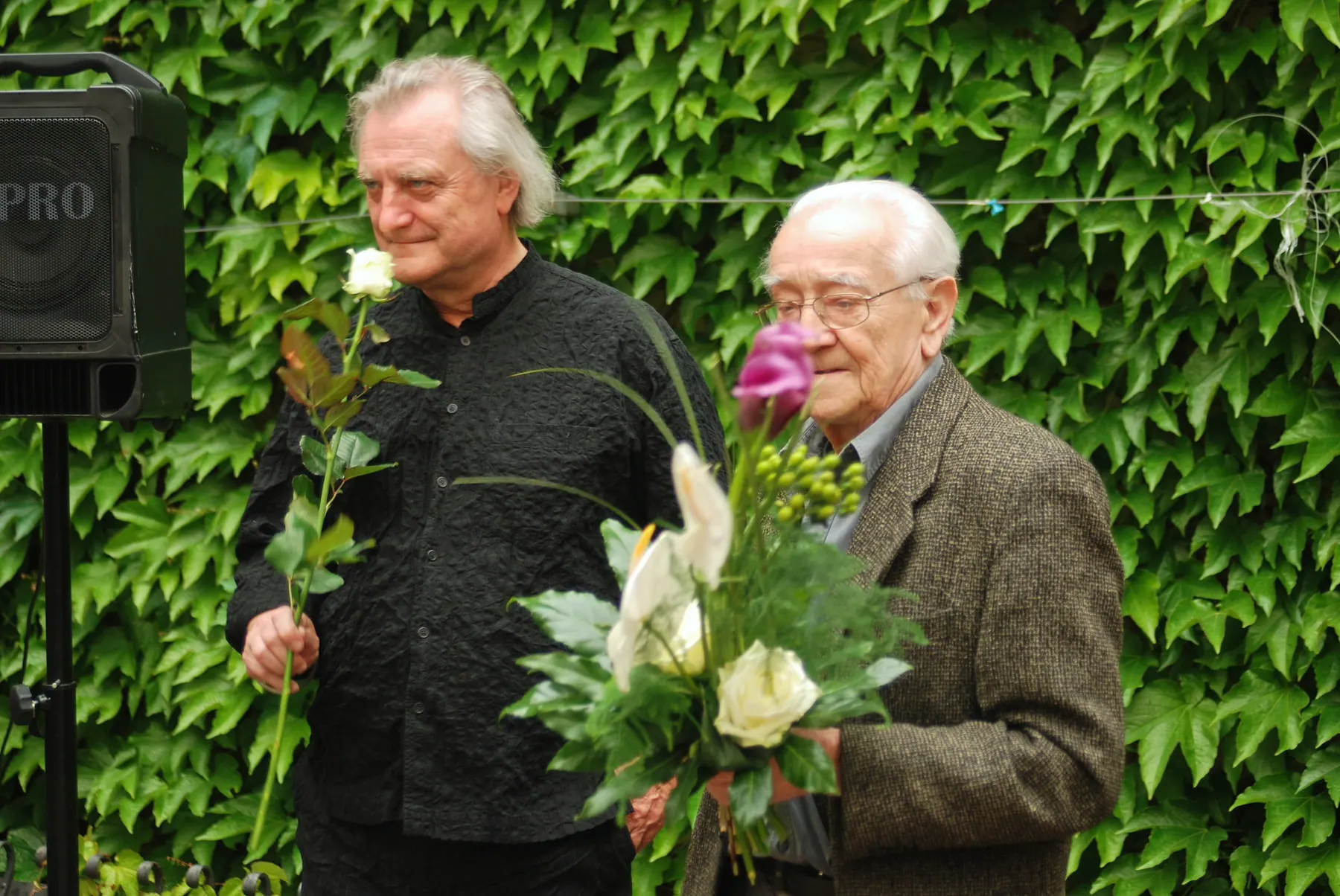 Two men holding a white rose and a bouquet of flowers in front of an ivy-covered wall
