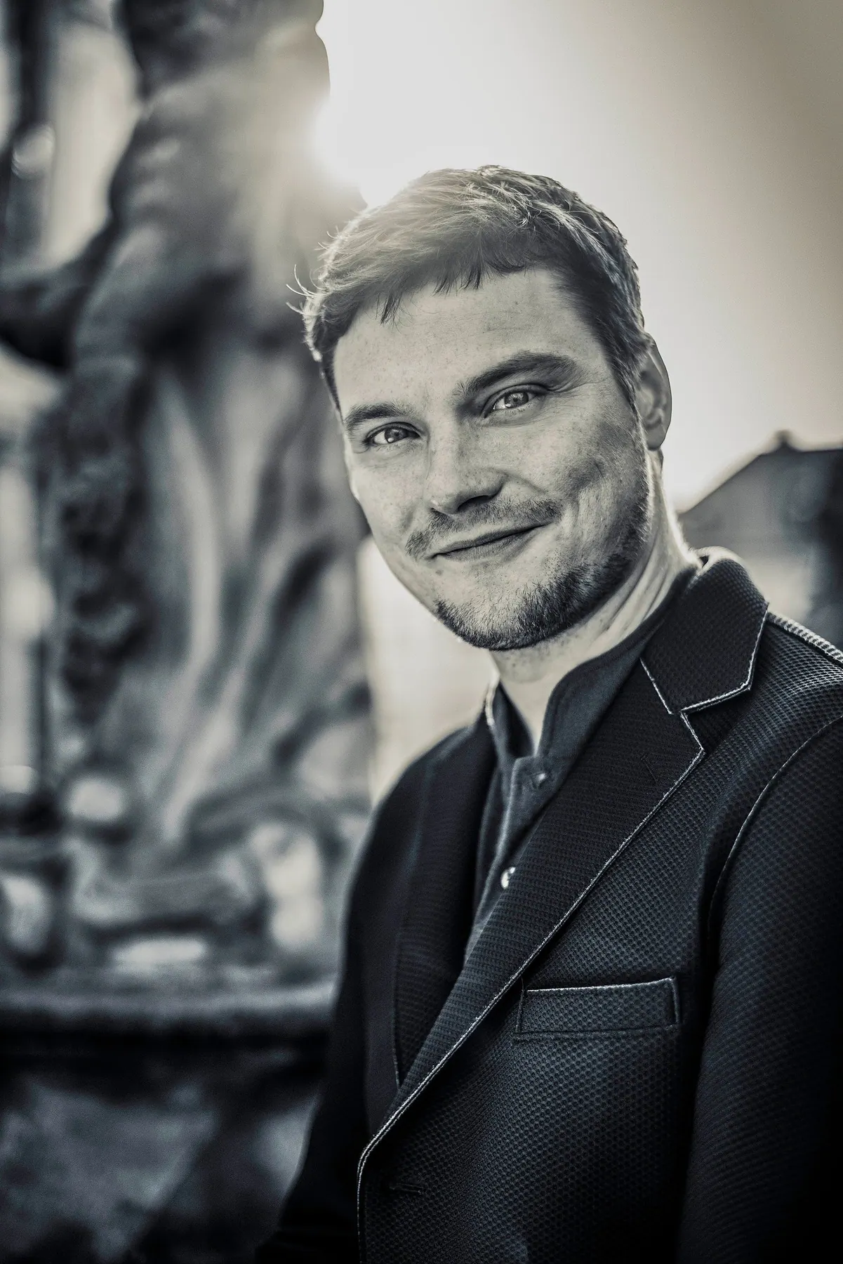 Black-and-white portrait of Milan Šípek smiling, with a stone statue behind him