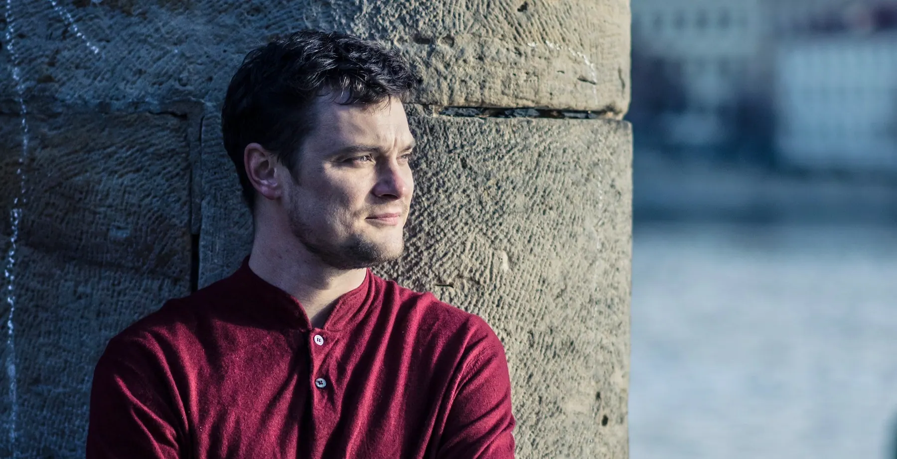 Milan Šípek in a red shirt leaning against a stone wall by the water