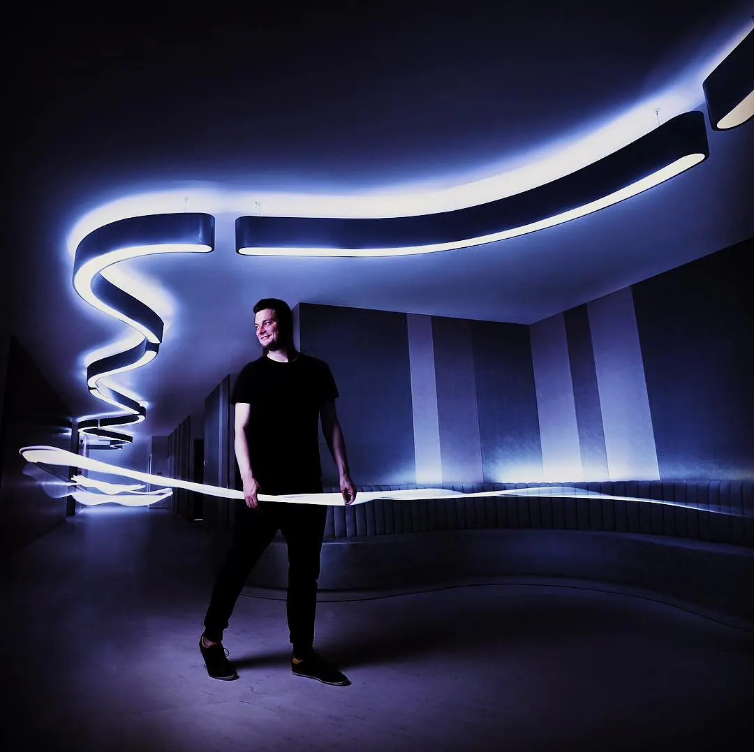 Milan Šípek standing beneath a glowing ribbon-shaped light installation in a dark room