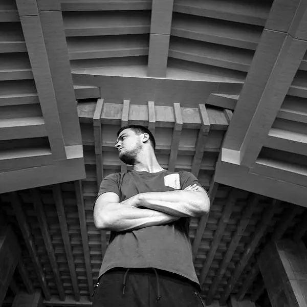 Black-and-white low-angle shot of Milan Šípek with arms crossed beneath a beamed ceiling
