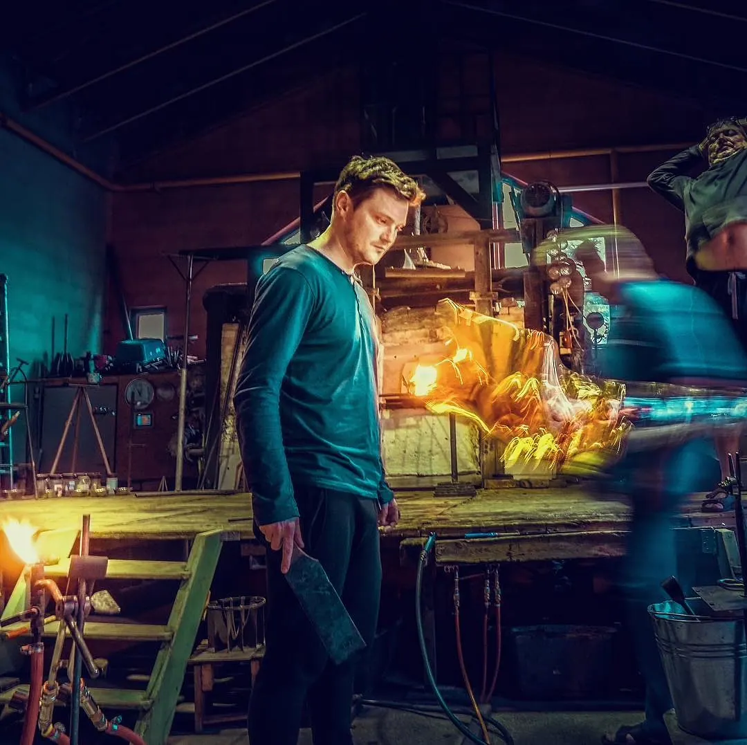 Milan Šípek in a glassworks beside a glowing furnace, with blurred workers behind him