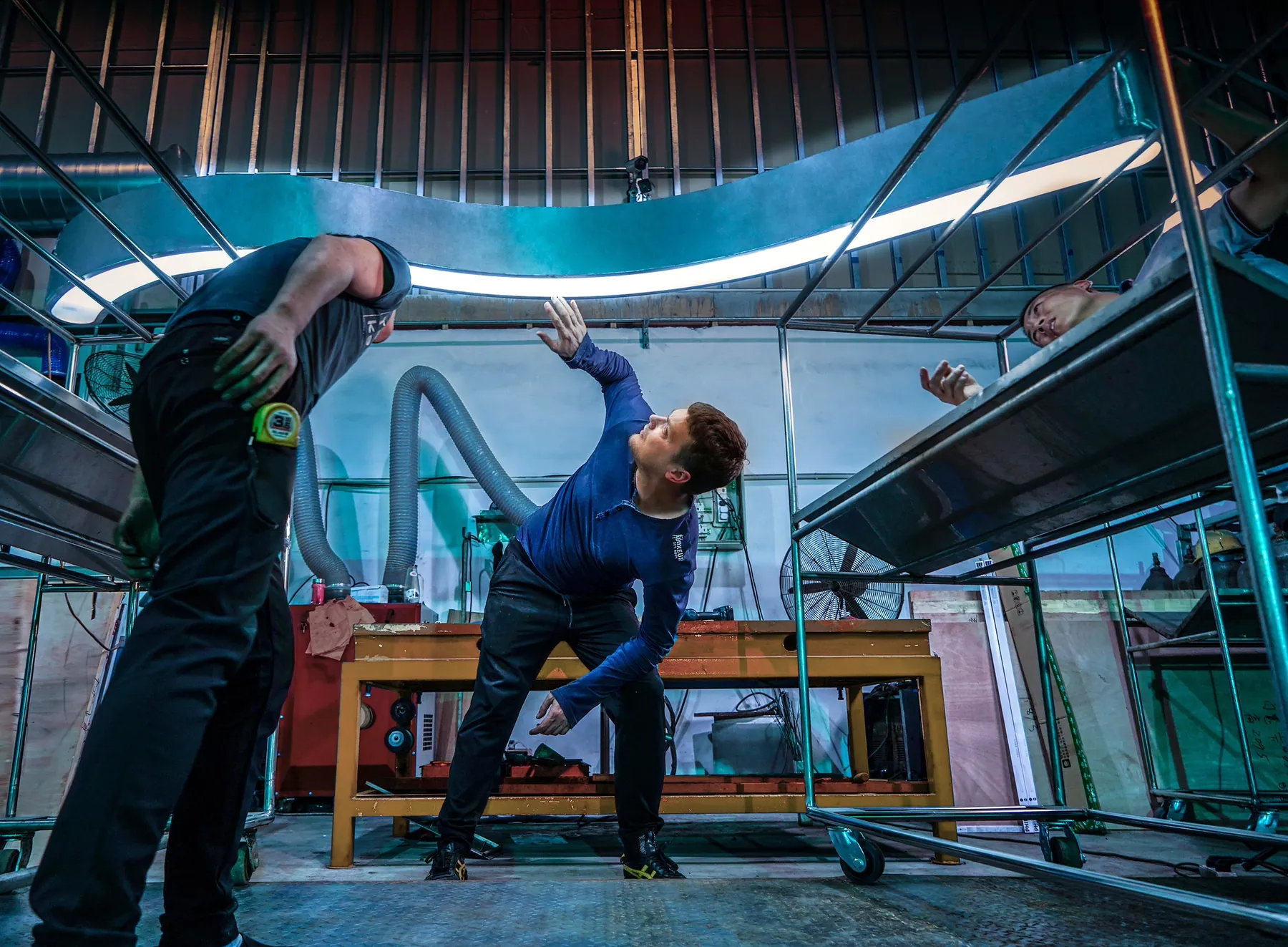 Men assembling a large curved light fixture on steel scaffolding in a workshop