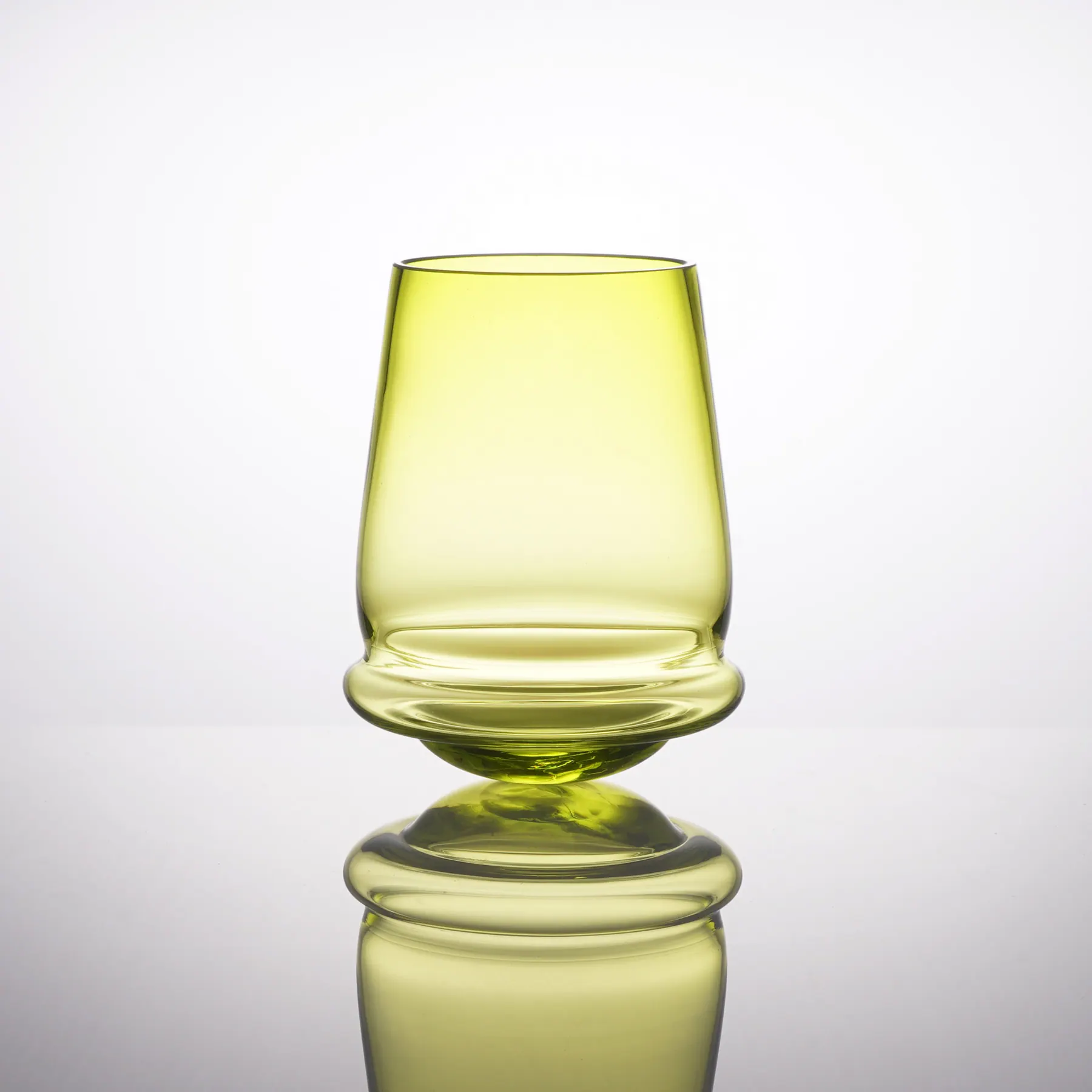 Upright yellow-green glass on its rounded base, reflected below, Tipsy glass collection