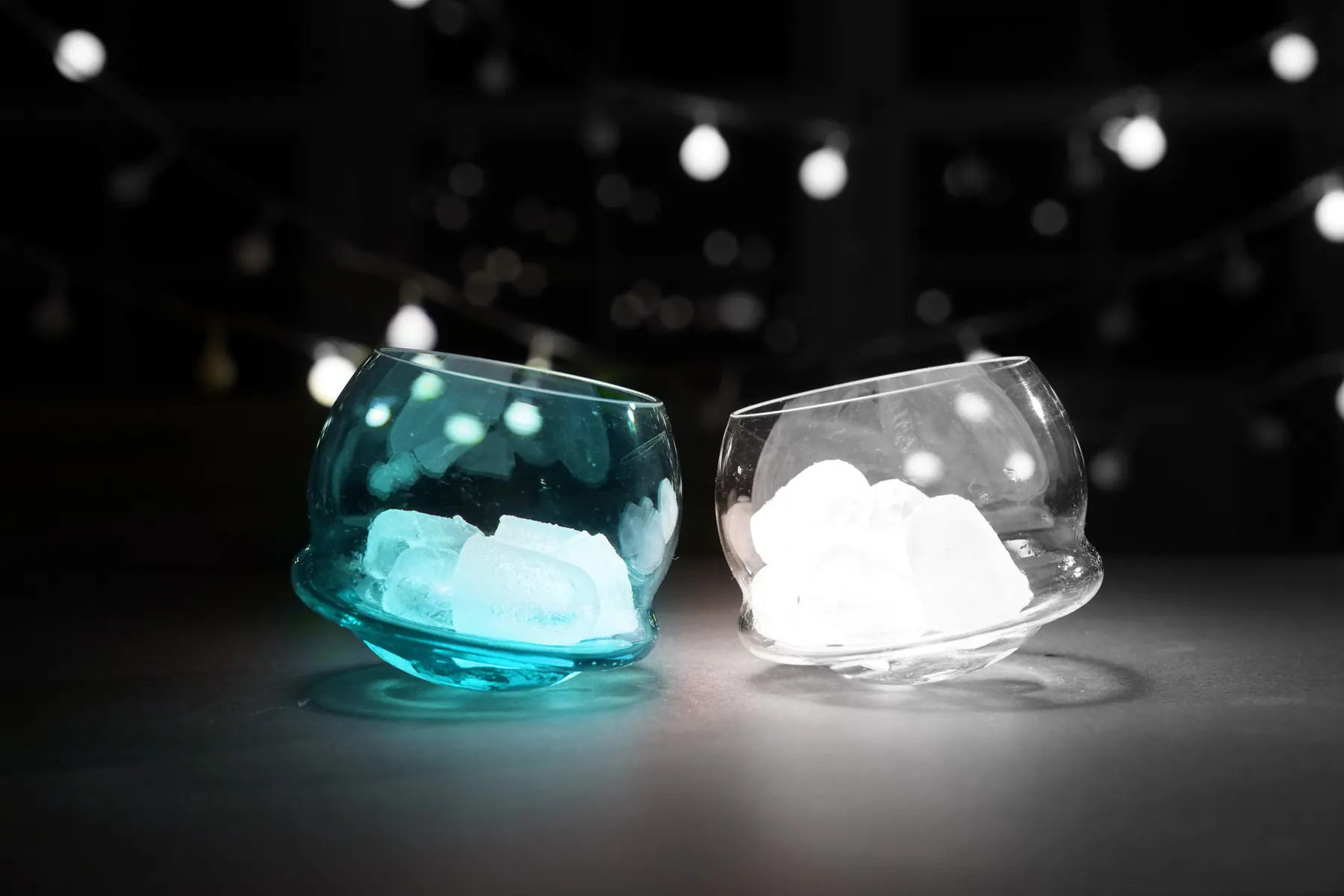 Turquoise and clear glasses with ice under string lights in the dark, Tipsy glass collection